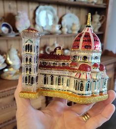 a hand holding up a christmas ornament in front of a shelf filled with china