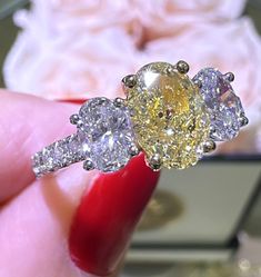 a yellow diamond ring sitting on top of a red manicured finger in front of a white rose