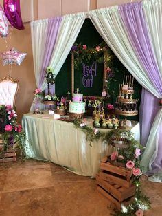 a table topped with lots of cakes and desserts next to purple draping