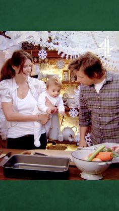 a man and woman holding a baby in front of a counter with food on it