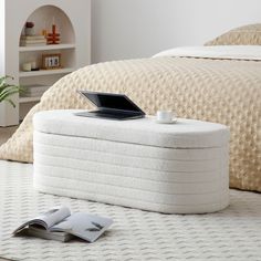 an open laptop computer sitting on top of a white ottoman next to a bed with beige bedspread