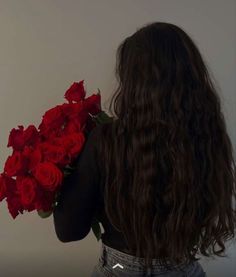 a woman with long dark hair holding red roses in her hand and looking at the wall