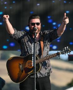 a man holding a guitar and giving the peace sign with his right hand while standing in front of a microphone