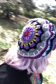 a woman with white hair wearing a crocheted hat and black jacket in the woods