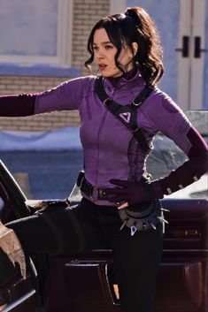 a woman in purple shirt and black pants standing next to a car with her hand on the door handle