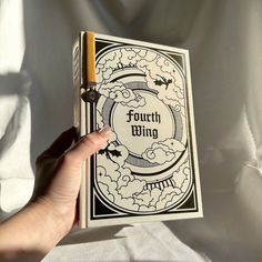 a hand holding an open book with the title fourth wing written in black and white