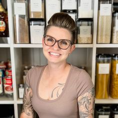 a woman with tattoos and glasses standing in front of a shelf full of spices, condiments and jars