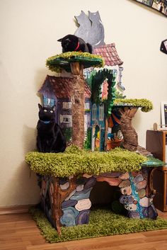 two black cats sitting on top of a house made out of fake grass and rocks