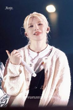 a woman with blonde hair wearing a white cardigan and holding her finger up in the air