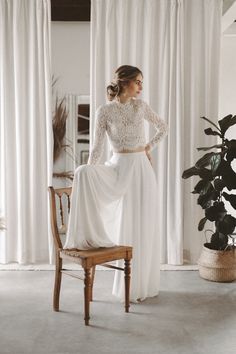 a woman standing in front of a window wearing a long sleeved wedding dress with an open back