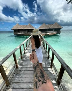 a man and woman holding hands while standing on a wooden bridge over water with thatched huts in the background