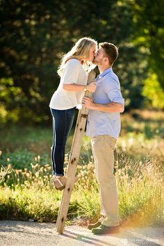 a man and woman kissing while standing on a ladder in front of some grass with trees behind them