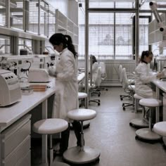 two women in white lab coats are working on microscopes and writing down notes while another woman stands at the counter