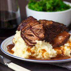 a white plate topped with mashed potatoes covered in meat and gravy next to a bowl of greens