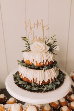 a three tiered cake with white icing and greenery on top, surrounded by cupcakes