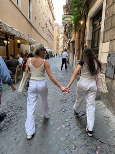 two women are holding hands while walking down the street in front of some people and buildings