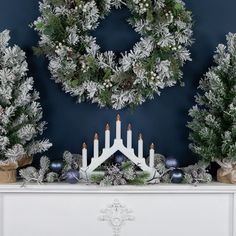 a white mantle with candles and christmas wreaths on it in front of a blue wall