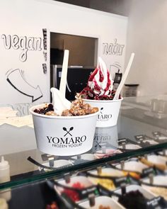 two ice cream bowls with spoons in them sitting on a glass counter top next to other desserts