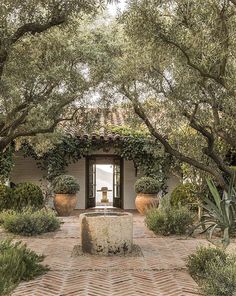 an outdoor fountain surrounded by trees and plants in front of a small building with tiled flooring