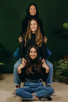 three women sitting on top of each other in front of a green wall and smiling