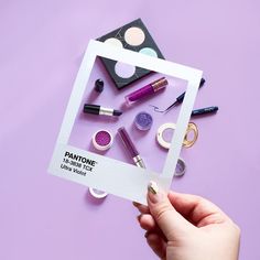 a person holding up a card with various makeup items in it on a purple surface