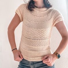 a woman standing in front of a white wall wearing a sweater and jeans with her hands on her hips