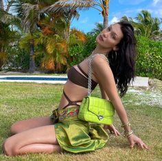 a woman sitting in the grass with a green purse on her shoulder and palm trees behind her