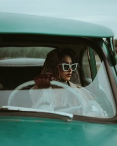 a woman sitting in the driver's seat of a green car wearing white sunglasses