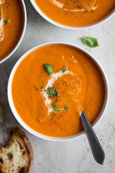 three bowls of tomato soup with basil and parmesan cheese on the top, next to bread