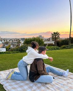 a man and woman sitting on top of a blanket in front of the ocean at sunset