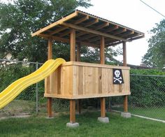 a wooden structure with a yellow slide in the grass next to a chain link fence