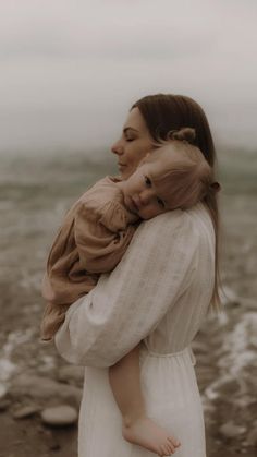 a woman holding a baby in her arms while standing on a rocky beach with the ocean in the background