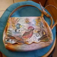 "Anuschka is the only family owned, designed, and ethically produced hand painted brand. Created with original artwork on finest ethically produced leathers, and unrivaled quality at an incredible value. This older vintage Anushka Purse is an ocean reef  scene with fish, coral, octopus, shells. 1980s. The leather is dyed a rainbow of colors on a sand brown background. Brass hardware. In excellent condition, the painting is gorgeous. The lining is clean and beautiful. 12\" long. 9.5\" tall. Strap Vintage Hand Painted Rectangular Bag, Artsy Hand Painted Shoulder Bag For Daily Use, Artistic Hand-painted Satchel Shoulder Bag, Artistic Hand Painted Satchel Shoulder Bag, Artsy Hand Painted Shoulder Bag For Everyday, Artsy Hand Painted Rectangular Shoulder Bag, Hand Painted Leather Bag, Painted Leather Bag, Ocean Reef