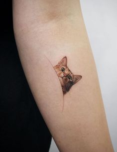 a small cat tattoo on the left inner arm and shoulder, with an orange kitten peeking out from behind it