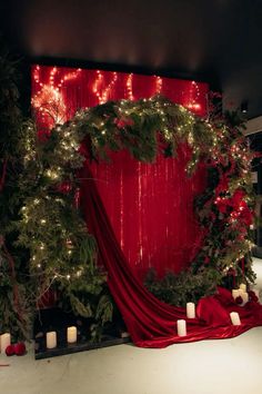 a red backdrop with candles and greenery on the floor next to a christmas wreath