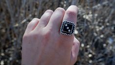 a person's hand with a black and white square ring on it, in front of some dry grass