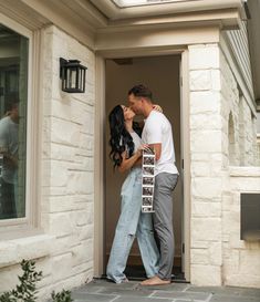 a man and woman kissing in front of a house with the door open to their side