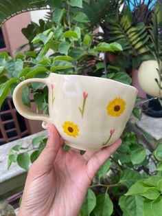 a hand holding up a cup with yellow flowers on it in front of some plants