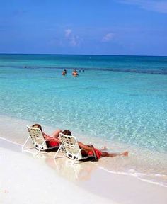 two people lounging on lounge chairs at the beach in front of clear blue water