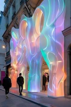 two people walking down the sidewalk in front of a building with colorful lights on it