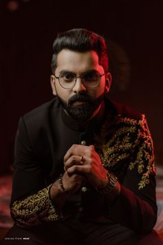 a man with glasses and a beard sitting in front of a dark background wearing a black suit