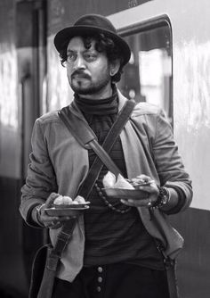 a man in a hat and scarf holding food on a plate while standing next to a train