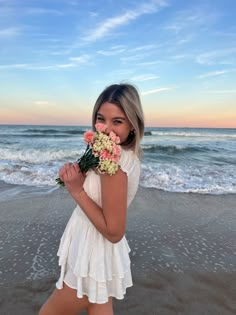 a woman standing on top of a beach next to the ocean with flowers in her hand