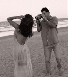 a man and woman standing on top of a beach next to the ocean taking pictures