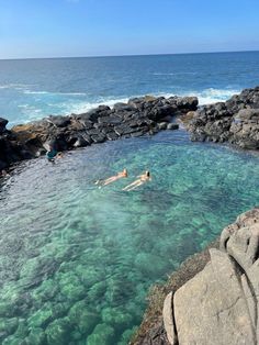 two people swimming in the ocean next to some rocks and water with clear blue skies