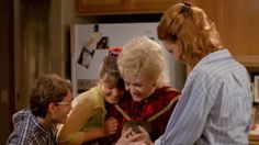 two women and three children standing in a kitchen looking at something on a cell phone