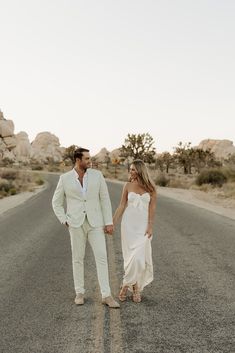 a man and woman walking down the middle of an empty road in white attire, holding hands