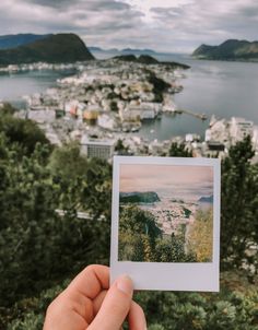 a person holding up a polaroid photo in front of a lake and cityscape