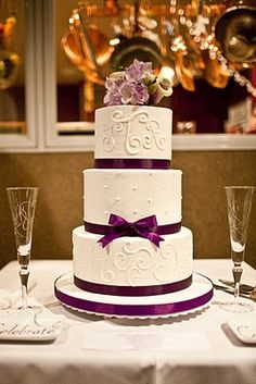 a three tiered wedding cake with purple ribbon and flowers on the top is surrounded by wine glasses