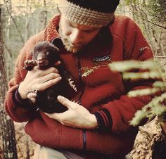 a man holding a small animal in his hands while wearing a hat and jacket with trees behind him